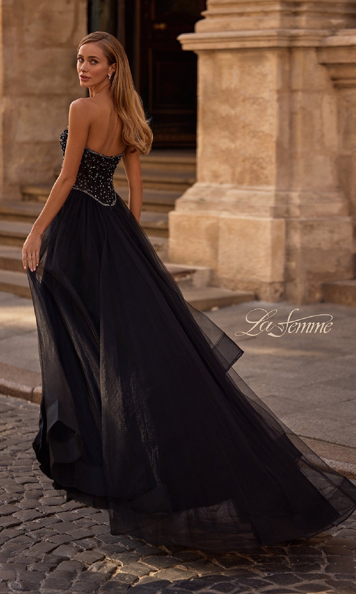 A woman in the La Femme 32856 strapless long black prom ball gown with a beaded bodice and tulle skirt walks on a cobblestone street in front of a stone building.